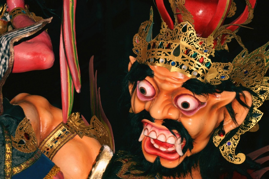 A dramatic, close-up photo of a colorful giant effigy mask known as an Ogoh-Ogoh. The effigy has a fearsome face with bulging eyes, white fangs, a black beard, and a traditional golden crown.