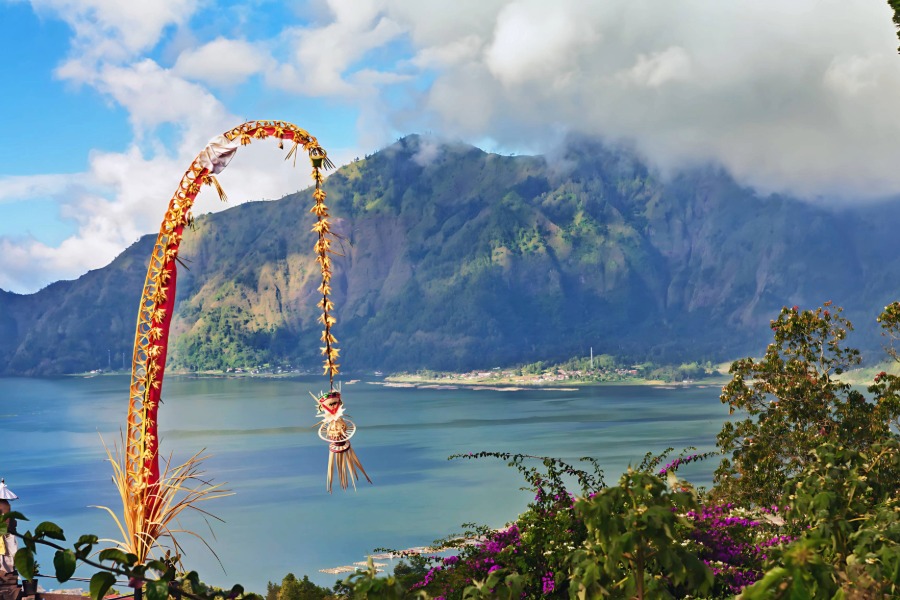 A beautiful scenic view of misty green hills and a blue lake (likely Lake Batur) in Bali, dominated by a tall, arching bamboo pole called a Penjor in the foreground