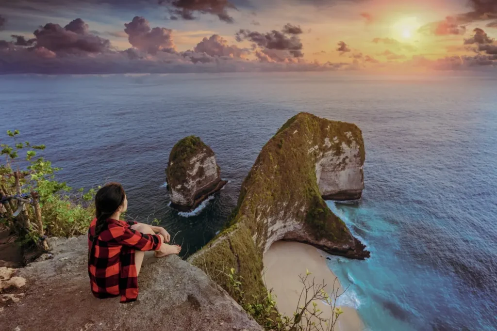 Person sits on cliff watching ocean and island rocks at sunset, showing slow travel in Bali.