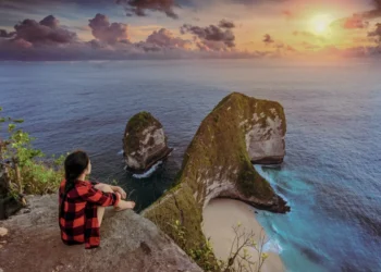 Person sits on cliff watching ocean and island rocks at sunset, showing slow travel in Bali.