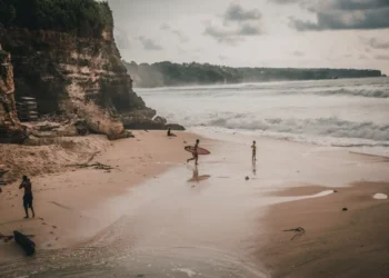 Surfers and visitors walk along Uluwatu Beach, surrounded by cliffs, waves, and a wide sandy shoreline.