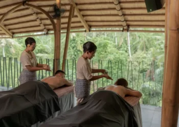 Two guests receive traditional Balinese massage while therapists prepare treatment in an open-air spa overlooking tropical greenery.