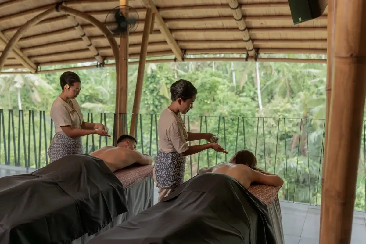 Two guests receive traditional Balinese massage while therapists prepare treatment in an open-air spa overlooking tropical greenery.