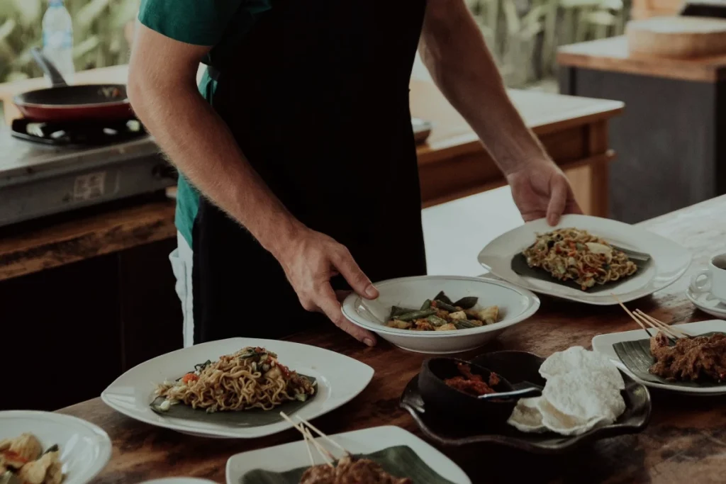 A cooking class instructor serves noodles, vegetables, satay, and other prepared dishes on a wooden table.