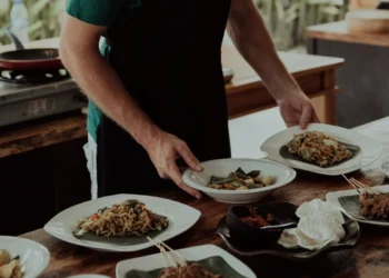 A cooking class instructor serves noodles, vegetables, satay, and other prepared dishes on a wooden table.