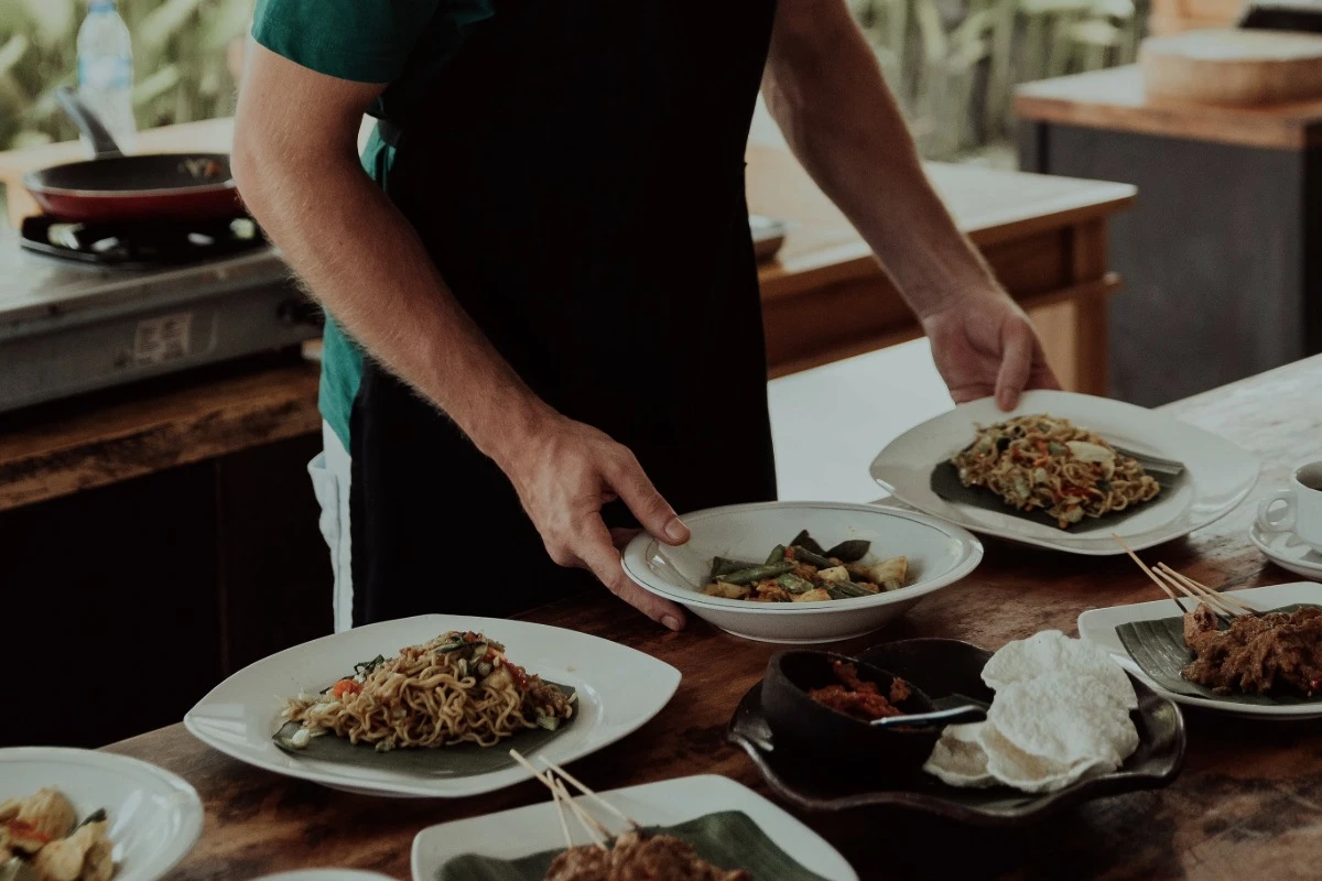 A cooking class instructor serves noodles, vegetables, satay, and other prepared dishes on a wooden table.