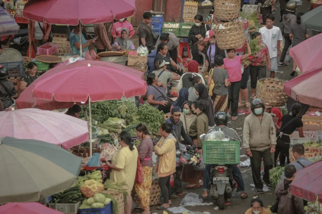 Crowded Badung Market with shoppers, vegetable stalls, motorbikes, and vendors carrying large woven baskets under colorful umbrellas.