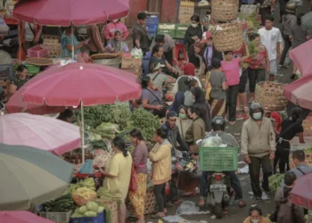 Crowded Badung Market with shoppers, vegetable stalls, motorbikes, and vendors carrying large woven baskets under colorful umbrellas.
