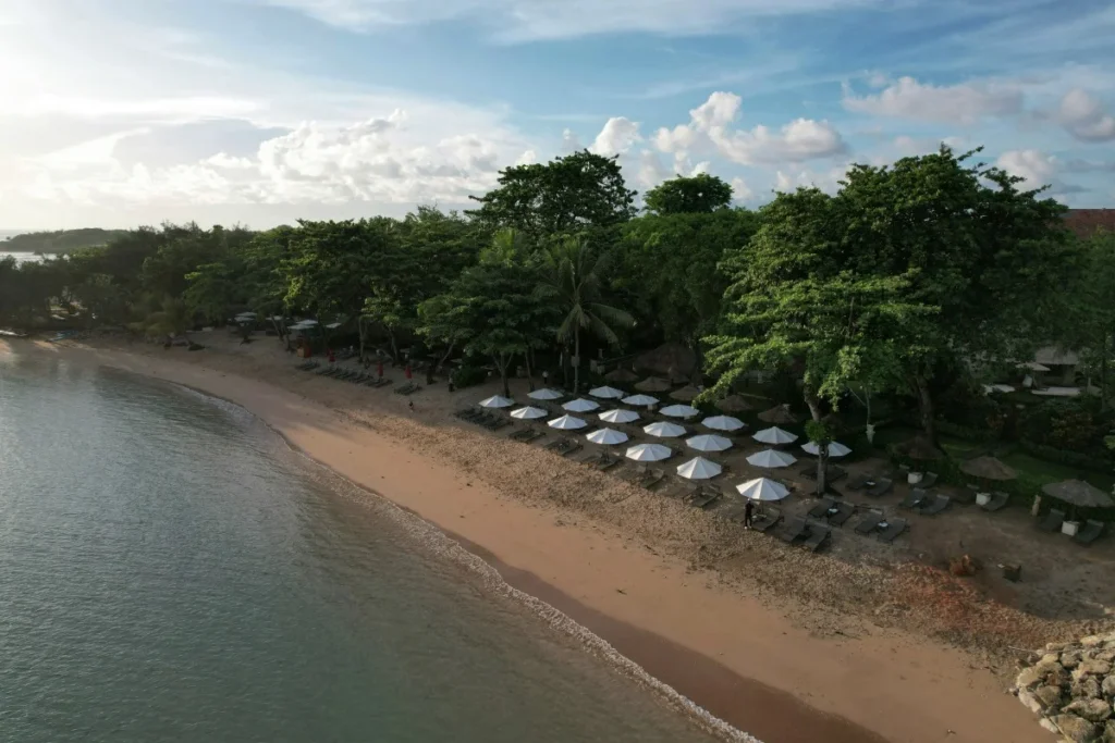 Aerial view of Nusa Dua beach with sandy shores, calm seawater, white umbrellas, sun loungers, and trees along the coast.