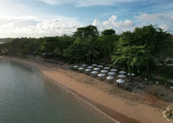 Aerial view of Nusa Dua beach with sandy shores, calm seawater, white umbrellas, sun loungers, and trees along the coast.