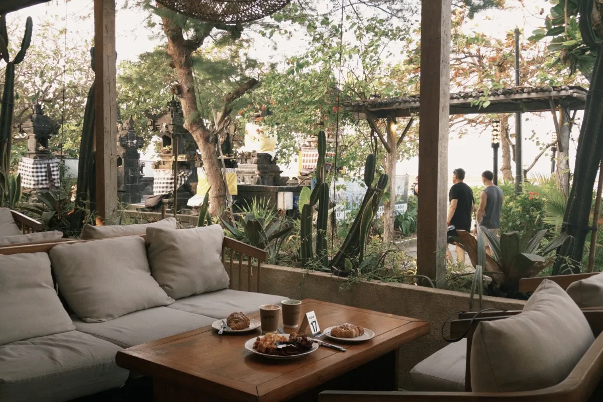 A quiet Sanur café with outdoor seating, soft cushions, coffee, and pastries served on a wooden table surrounded by greenery.