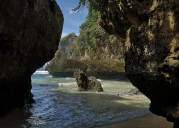 Ocean waves flow through a rocky cave entrance at Suluban Beach, with jagged cliffs and shallow water along the shore.