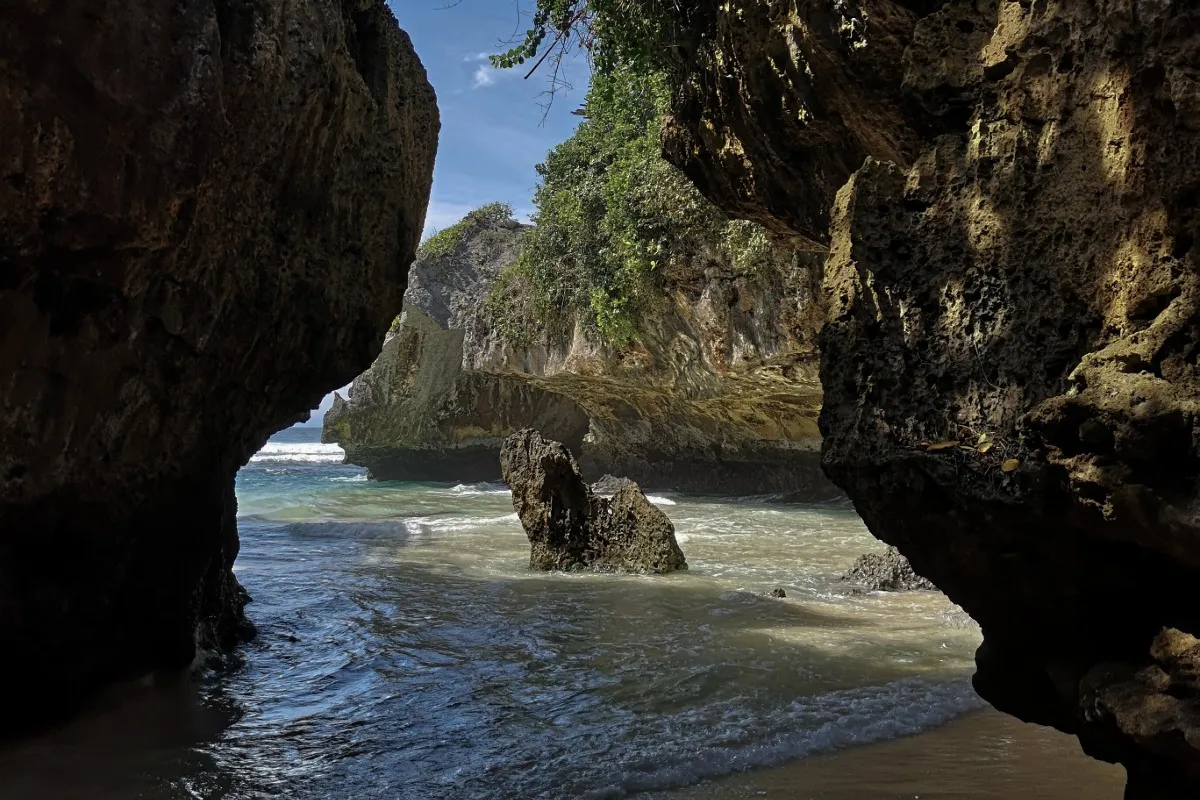 Ocean waves flow through a rocky cave entrance at Suluban Beach, with jagged cliffs and shallow water along the shore.