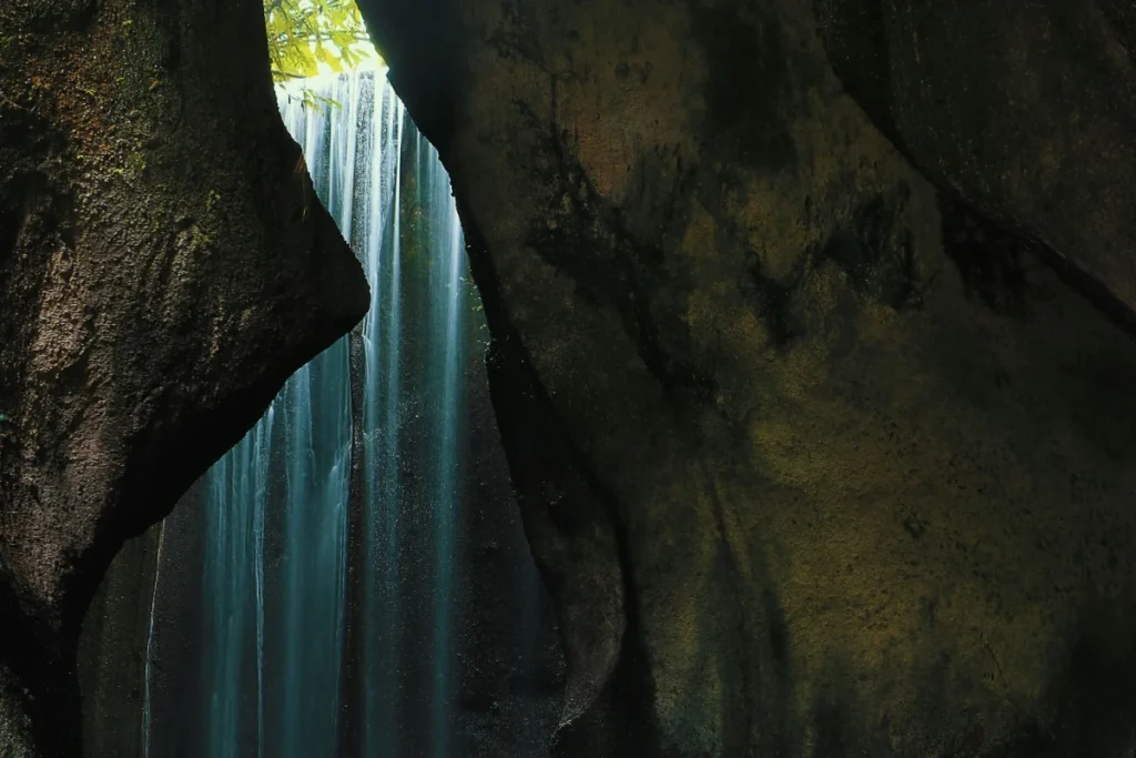 Tukad Cepung Waterfall flows through a narrow rock opening, with sunlight illuminating the falling water inside the cave.